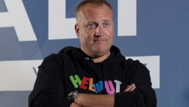 Man with blue eyes wearing a black hoodie with colorful text, arms crossed.