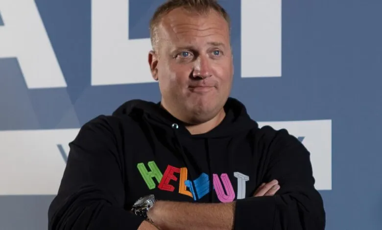 Man with blue eyes wearing a black hoodie with colorful text, arms crossed.