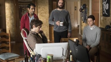 Four men in a cluttered room, two looking at computers, one holding a mug, and one standing.