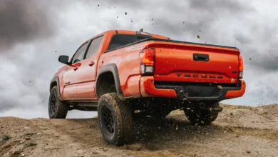 Orange Toyota Tacoma TRD Off-Road truck driving on a dirt trail, kicking up dust.