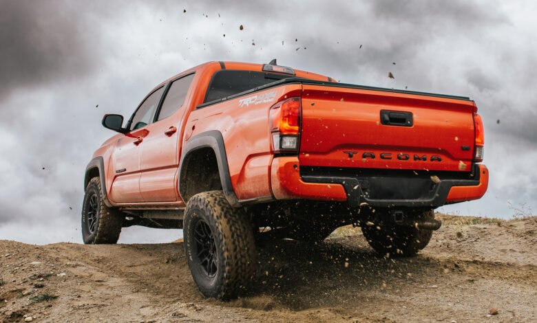 Orange Toyota Tacoma TRD Off-Road truck driving on a dirt trail, kicking up dust.