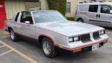 Silver 1980s Oldsmobile Cutlass Supreme coupe with red pinstripes parked outdoors.