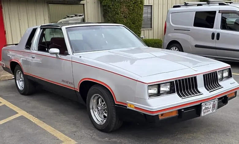 Silver 1980s Oldsmobile Cutlass Supreme coupe with red pinstripes parked outdoors.
