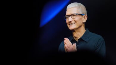 Tim Cook, CEO of Apple, smiles with hands clasped in front of a blue spotlight.