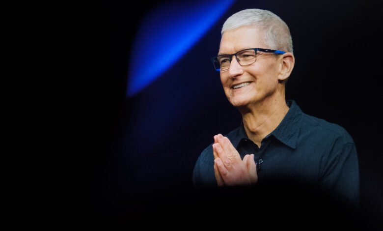 Tim Cook, CEO of Apple, smiles with hands clasped in front of a blue spotlight.