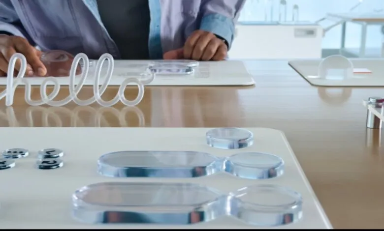 A person's hands interacting with clear, glass-like objects on a table, including a 'hello' sign.
