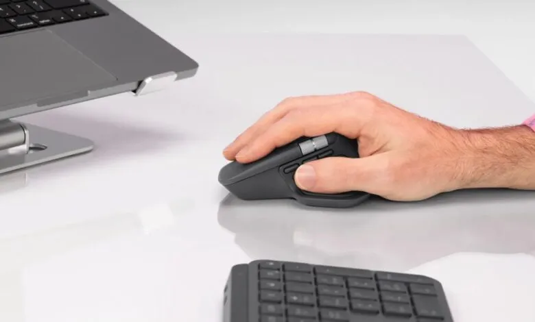 Close-up of a hand using a dark gray Logitech MX Master 4 mouse next to a laptop.