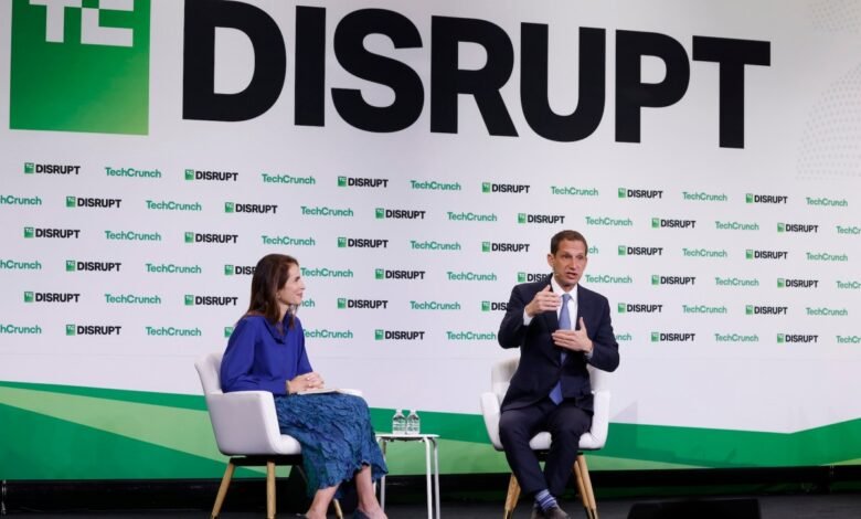Two speakers on stage at TechCrunch Disrupt, with a large "DISRUPT" logo behind them.