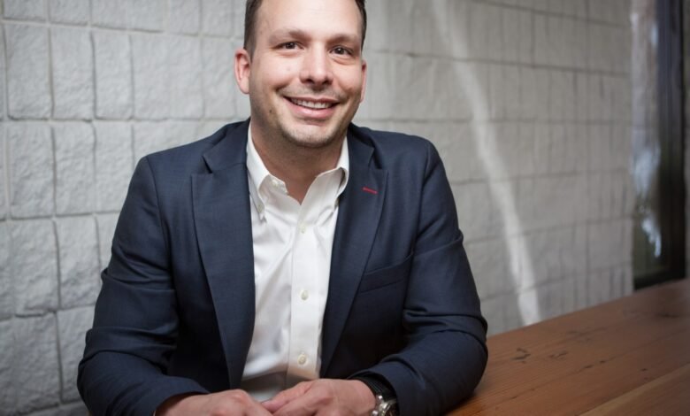 A smiling man in a dark suit and white shirt sits at a wooden table.