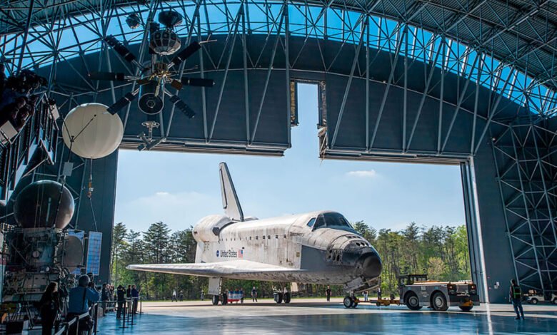 Space Shuttle Discovery inside a large hangar with other spacecraft and visitors.