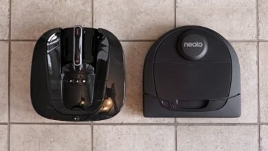 Two black robot vacuum cleaners, one with a handle and the other with a Neato logo, sit on a tiled floor.