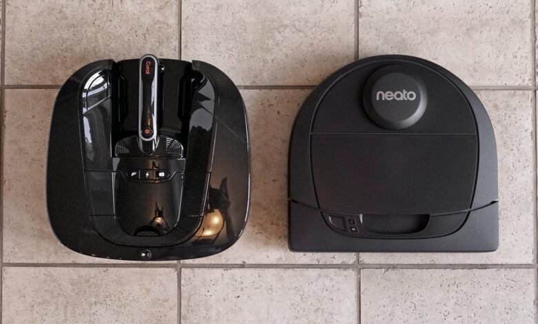 Two black robot vacuum cleaners, one with a handle and the other with a Neato logo, sit on a tiled floor.