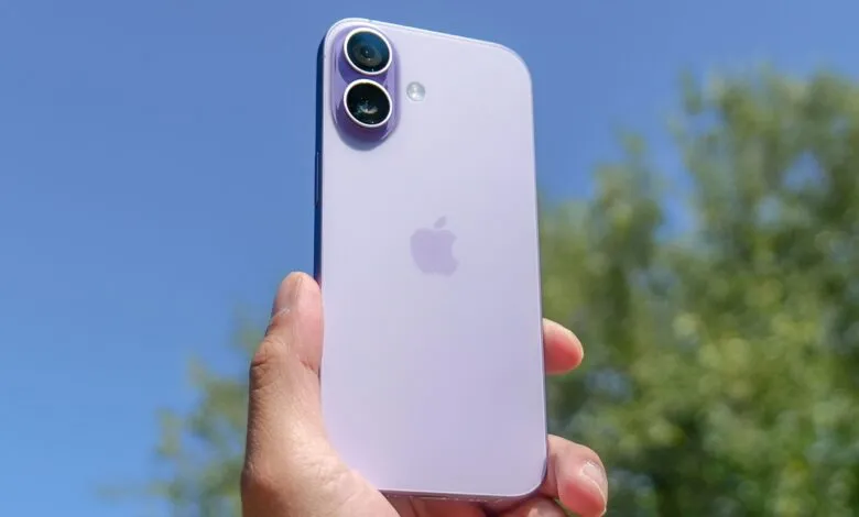 A hand holds a lavender iPhone 14 against a bright blue sky with blurred green trees.