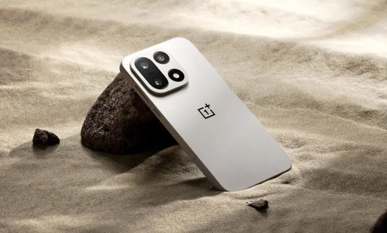 A light gray OnePlus smartphone with a prominent camera module rests on sand.