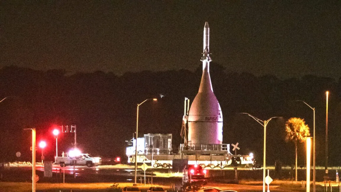 NASA rocket on transport crawler at night, illuminated by lights.