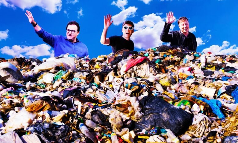 Three men waving from atop a large pile of trash under a blue sky.