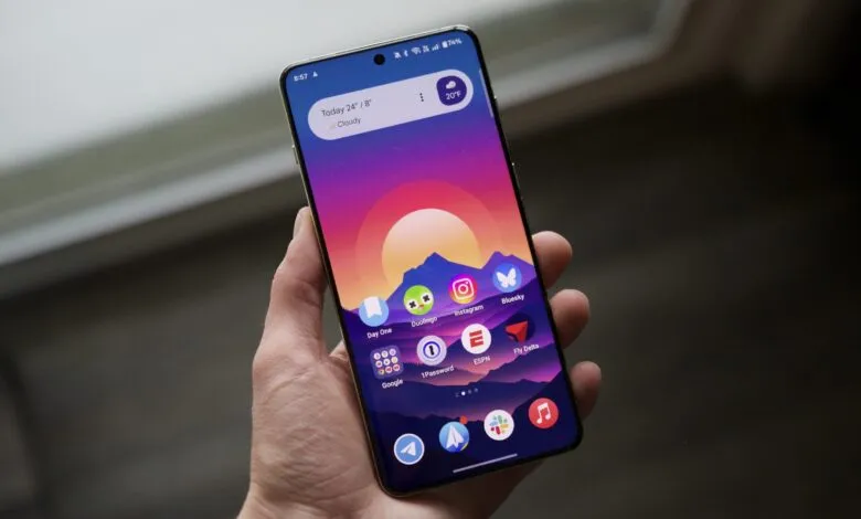 A person holds a smartphone displaying the OxygenOS 15 home screen with a vibrant mountain sunset wallpaper.