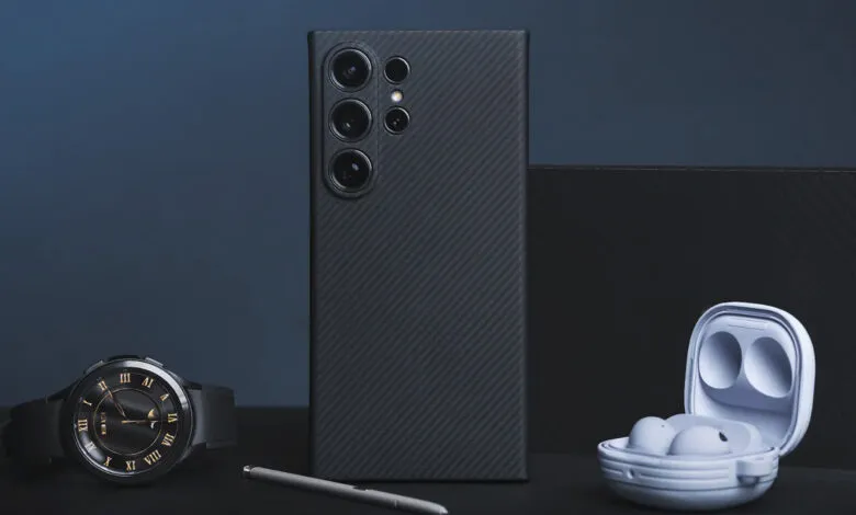 A dark grey smartphone with a carbon fiber case, a smartwatch, and wireless earbuds are arranged on a dark surface.