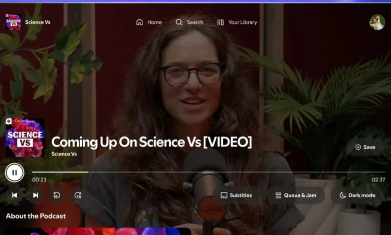 Woman with glasses speaking in front of a microphone, with podcast artwork and plants in the background.