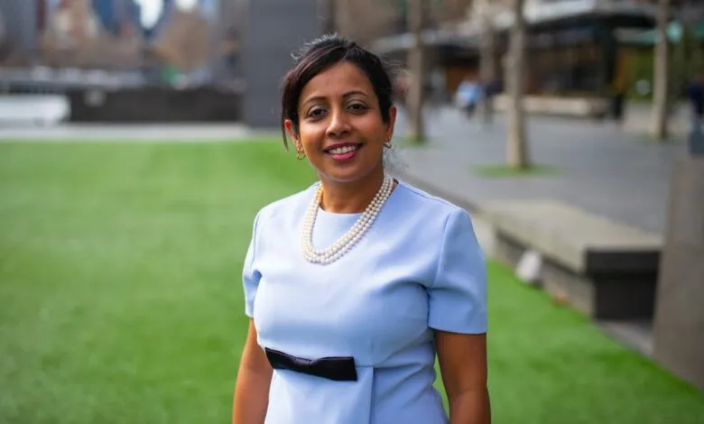 A woman in a light blue dress and pearl necklace smiles at the camera outdoors.