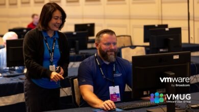 A woman in a blue shirt and black cardigan smiles while looking at a computer screen, with a man in a blue shirt seated at the same computer.