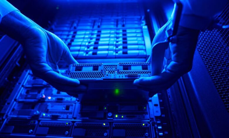 Hands installing a server component in a data center rack with blue lighting.