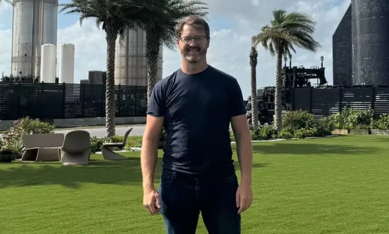 Man in dark t-shirt and jeans standing on a green lawn with palm trees and rocket structures.