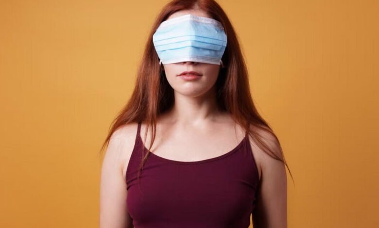 Woman with a blue surgical mask covering her eyes against an orange background.