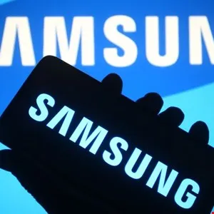 A hand holds a smartphone displaying the Samsung logo, with the Samsung logo visible on a blue screen in the background.