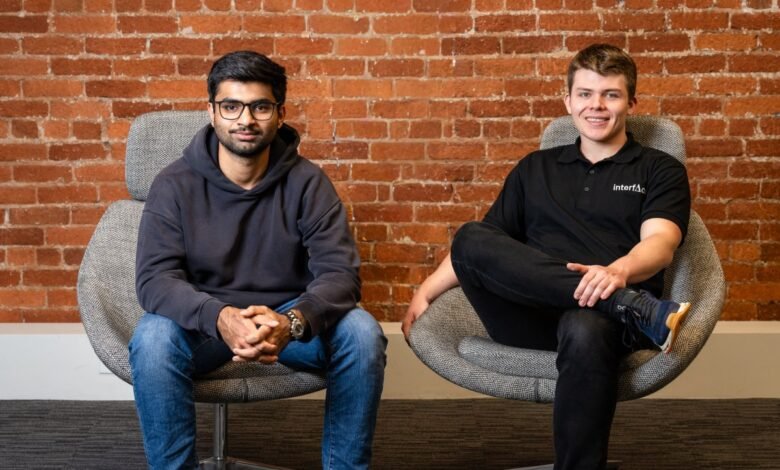 Two men sitting in chairs against a brick wall, one wearing glasses and a hoodie, the other in a polo shirt.
