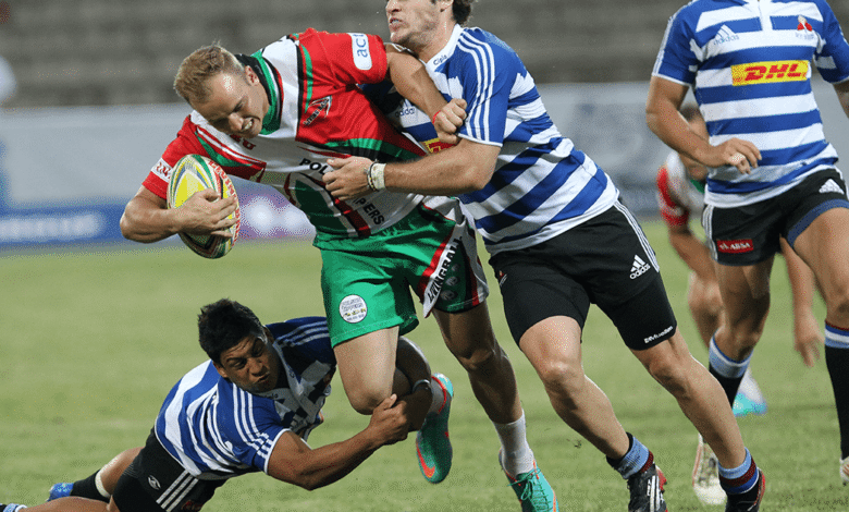 Rugby players in a tackle during a match, one player in red and green shorts is tackled by two opponents.
