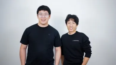 Two smiling Asian men pose against a white wall, one wearing glasses.