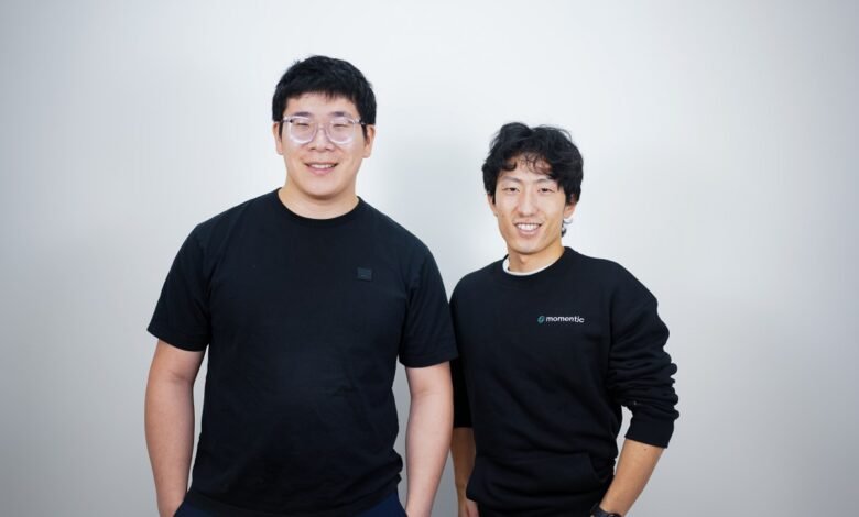Two smiling Asian men pose against a white wall, one wearing glasses.