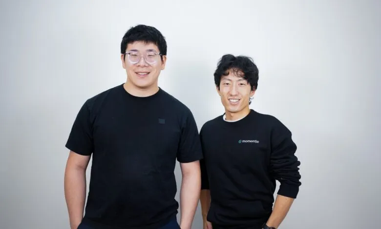Two smiling Asian men pose against a white wall, one wearing glasses.