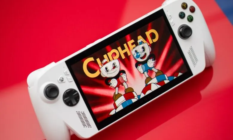 White handheld gaming console displaying the Cuphead game on a red surface.