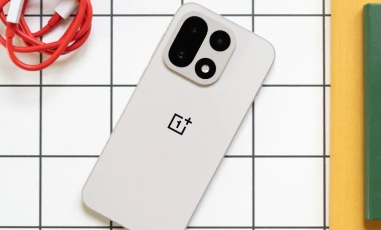 A light gray OnePlus smartphone with a prominent camera module and logo, next to red charging cables and books.