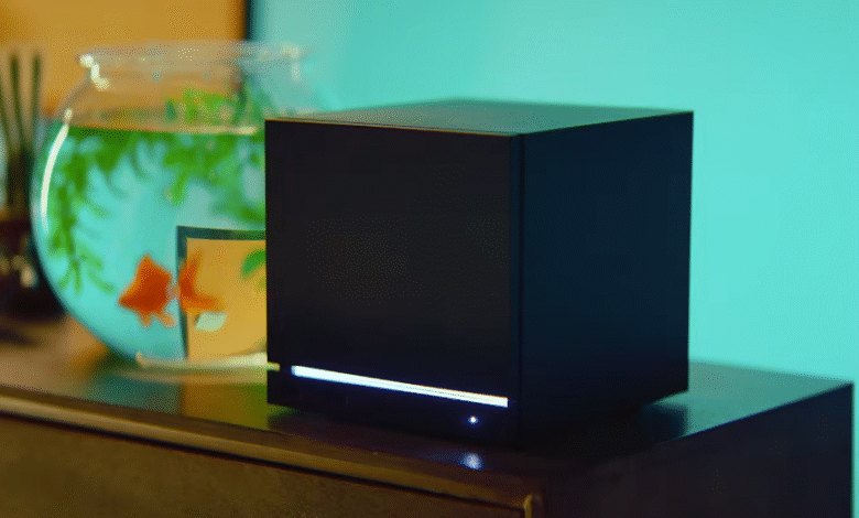 A black cubic device with a glowing blue light sits on a dark surface next to a fishbowl.