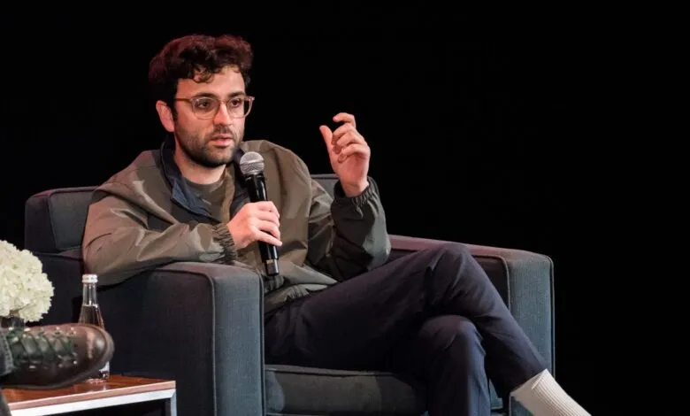 Man with glasses and curly hair speaking into a microphone while seated on a dark gray armchair.