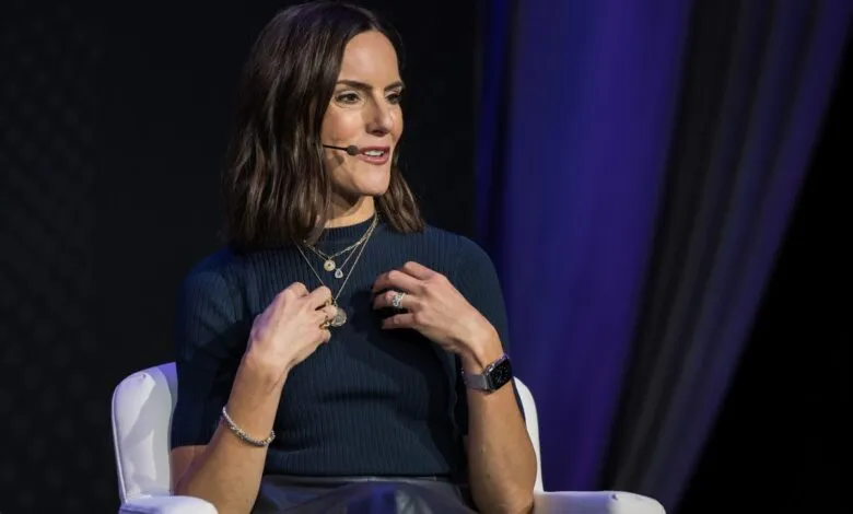 Woman with dark hair wearing a navy blue top and multiple necklaces, speaking at an event.