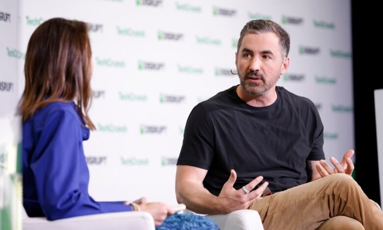 Man in black t-shirt and tan pants speaking animatedly, with a woman in a blue shirt seated opposite him.