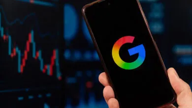 A hand holds a smartphone displaying the Google logo against a blurred stock market chart.