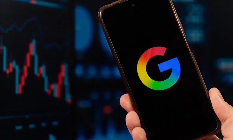 A hand holds a smartphone displaying the Google logo against a blurred stock market chart.