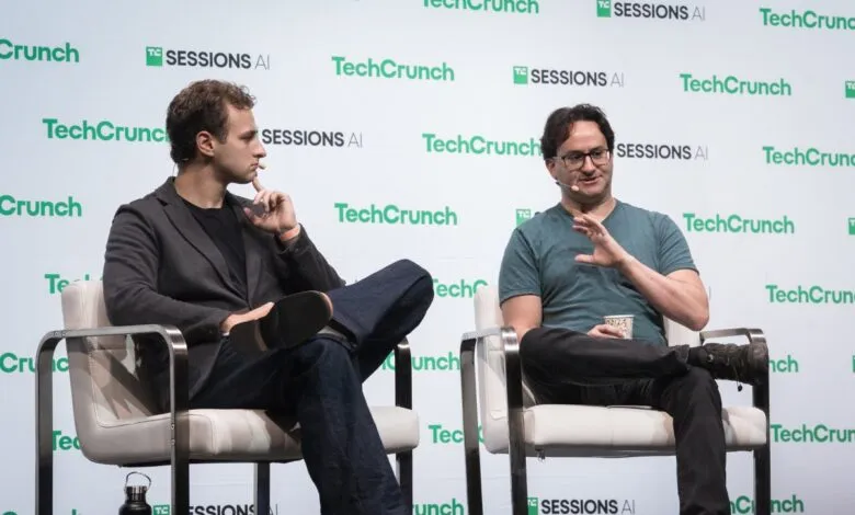Two men sit on chairs on a stage with a TechCrunch backdrop.