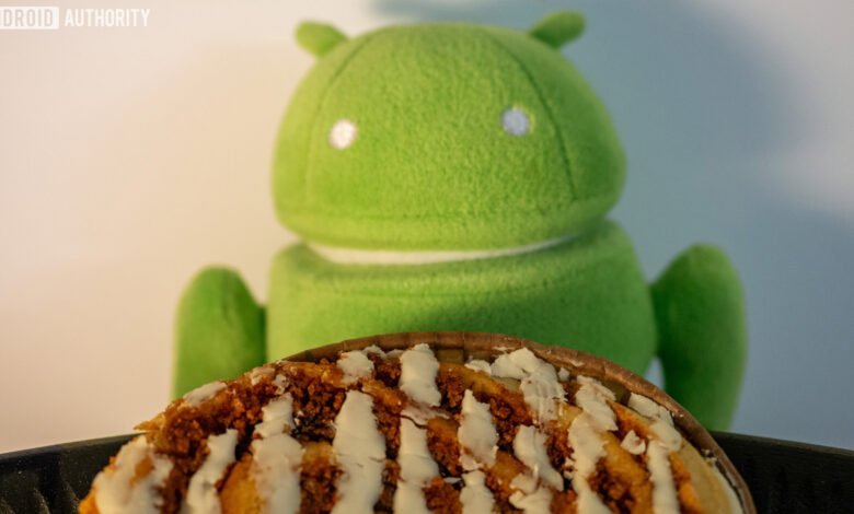 A green Android robot plush toy sits behind a cinnamon bun with white icing.