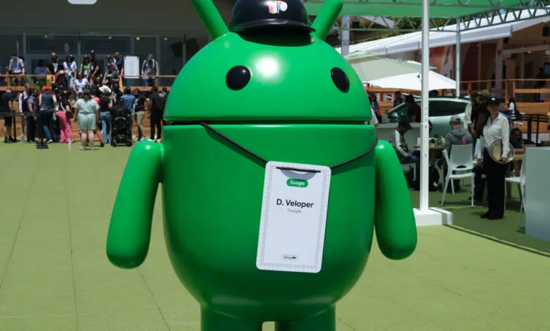 A large, green Android robot mascot wearing a black hat and a Google developer badge.