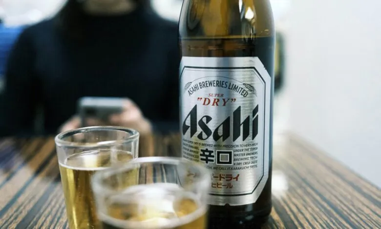 Asahi beer bottle and two glasses on a wooden table, person using phone in background.