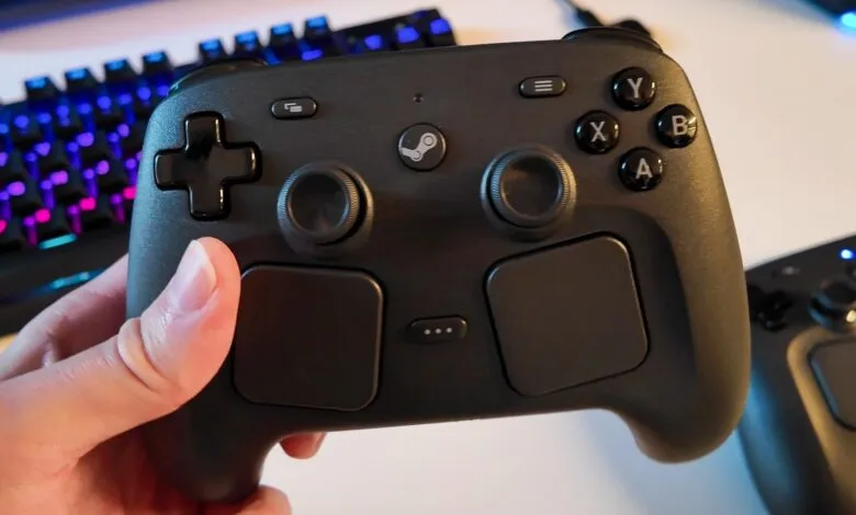 Close-up of a black Steam controller held in a hand, with a colorful keyboard in the background.