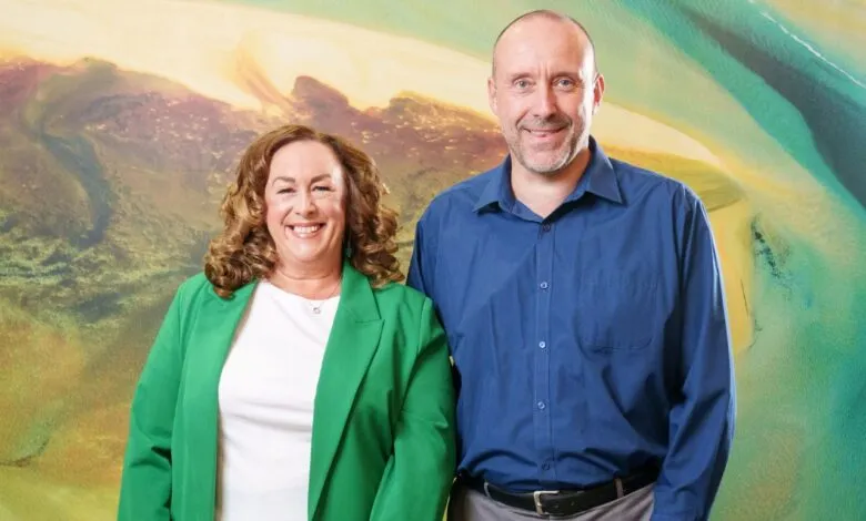 A smiling woman in a green blazer and a man in a blue shirt stand together.