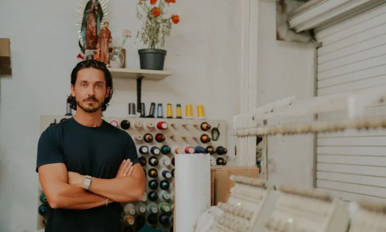 Man with arms crossed in a workshop with spools of thread and embroidery machines.