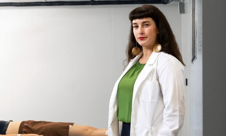 Woman in lab coat and green shirt with large earrings stands next to a person lying down.